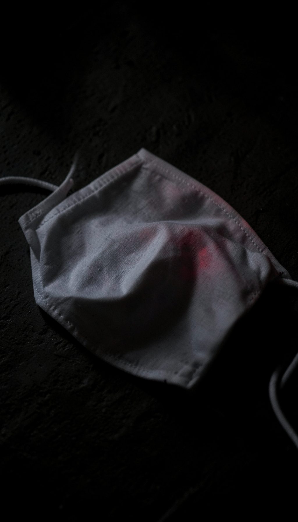 Dark photograph of a neatly folded white cotton face mask on a textured surface, deep shadow and minimal lighting