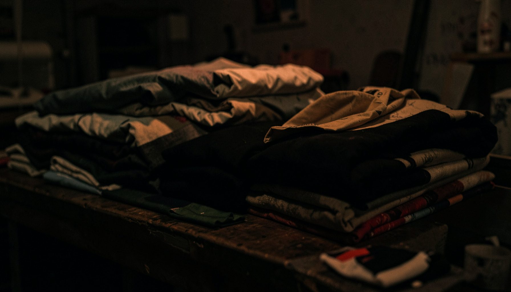 Dark moody photograph of folded cotton garments stacked on a textile workshop table, deep shadows and warm muted tones