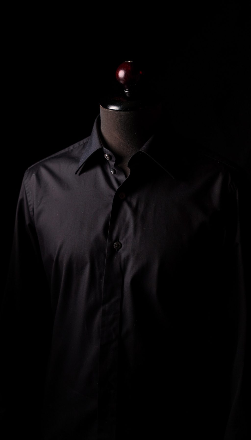 Dark photograph of a formal cotton shirt on a tailor's form, crisp collar detail, deep shadow background