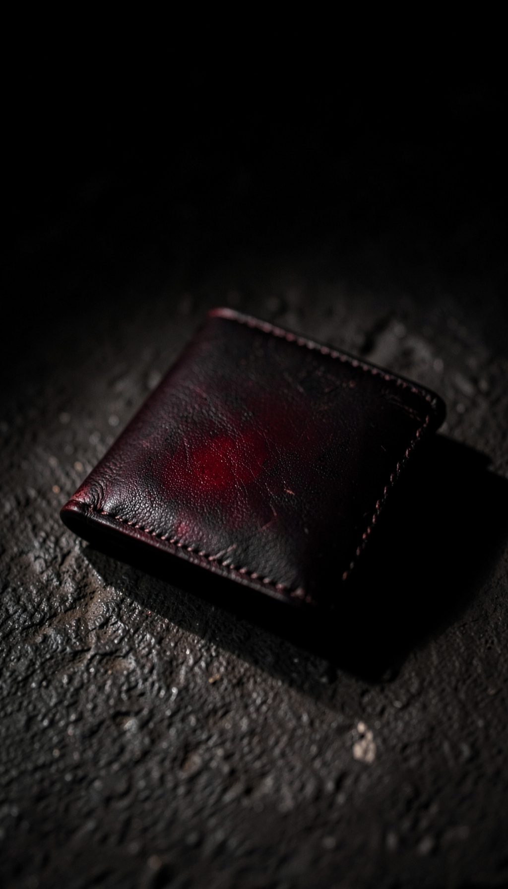 Dark moody photograph of a leather wallet resting on a textured surface, subtle highlights on stitching