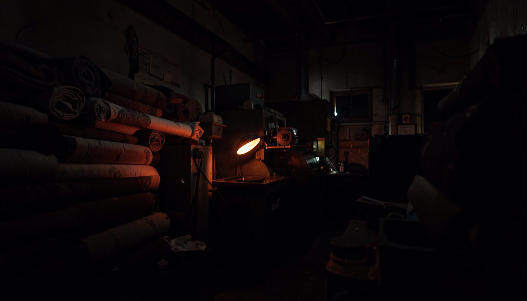 Dark industrial textile workspace with rolls of fabric stacked in shadow, warm accent lighting on machinery