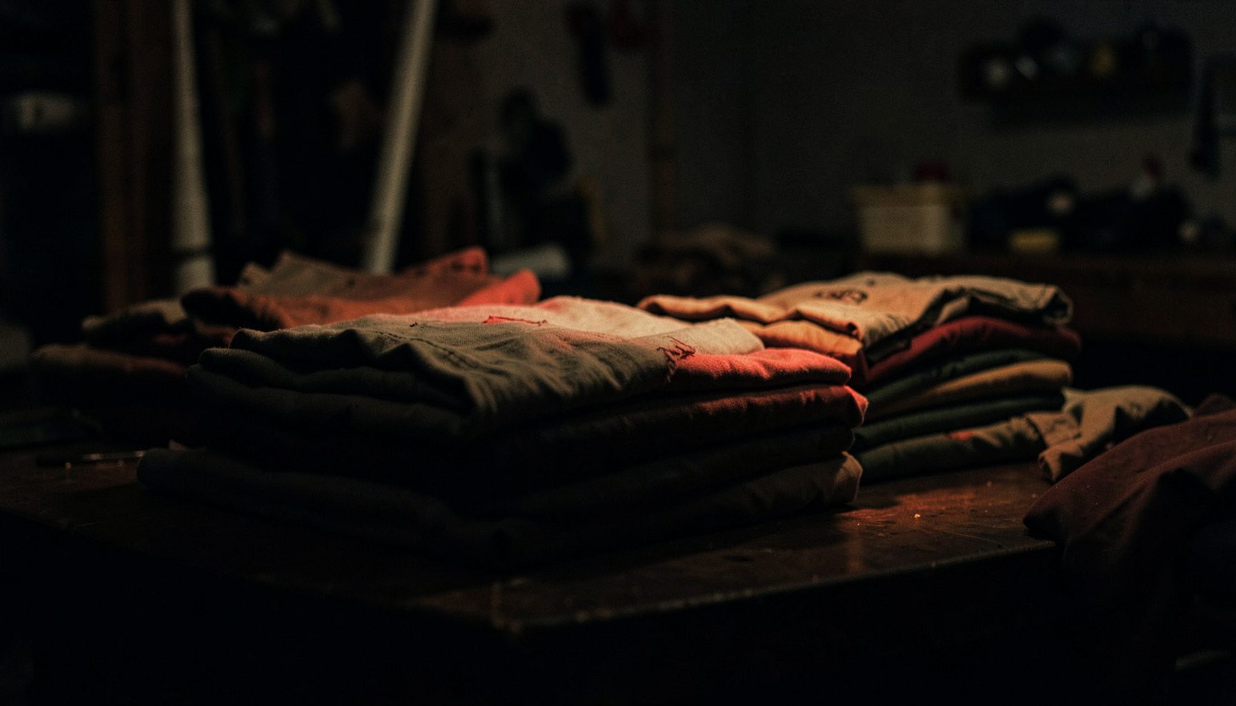 Dark photograph of neatly folded cotton garments stacked on a workshop table, deep shadows and warm muted tones