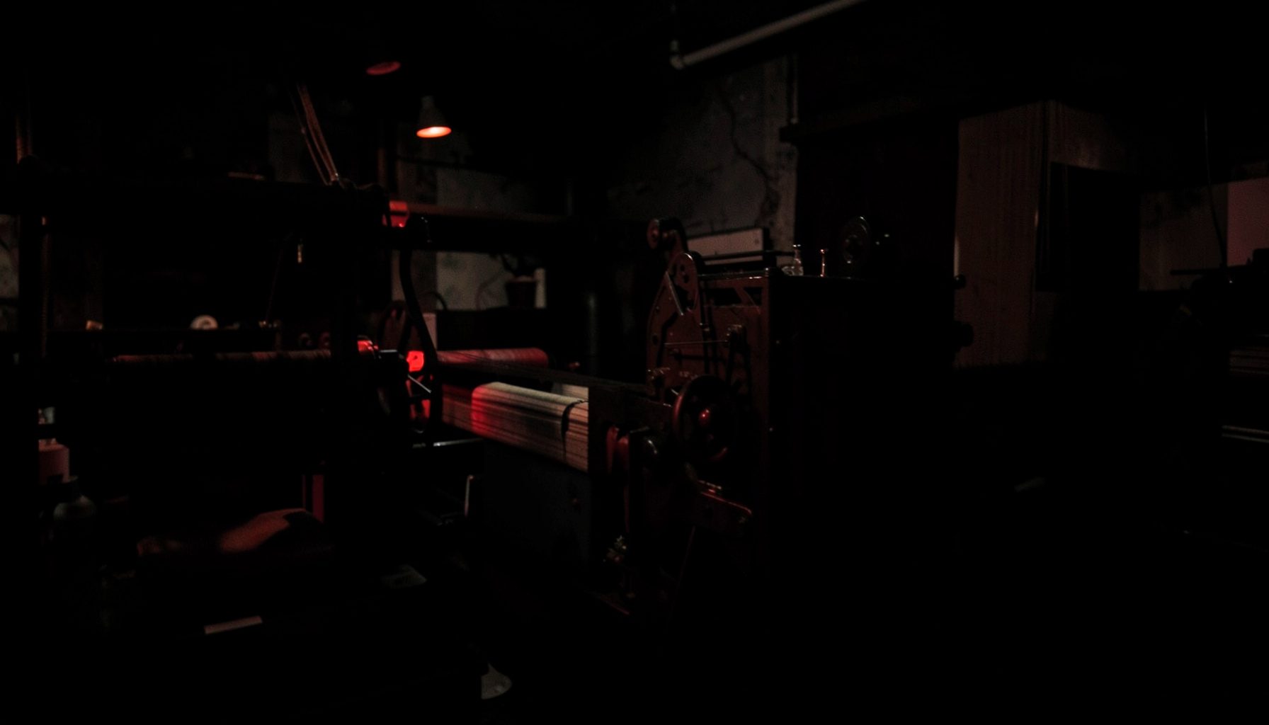 Dark atmospheric photograph of textile machinery in low light, deep blacks and muted red accents