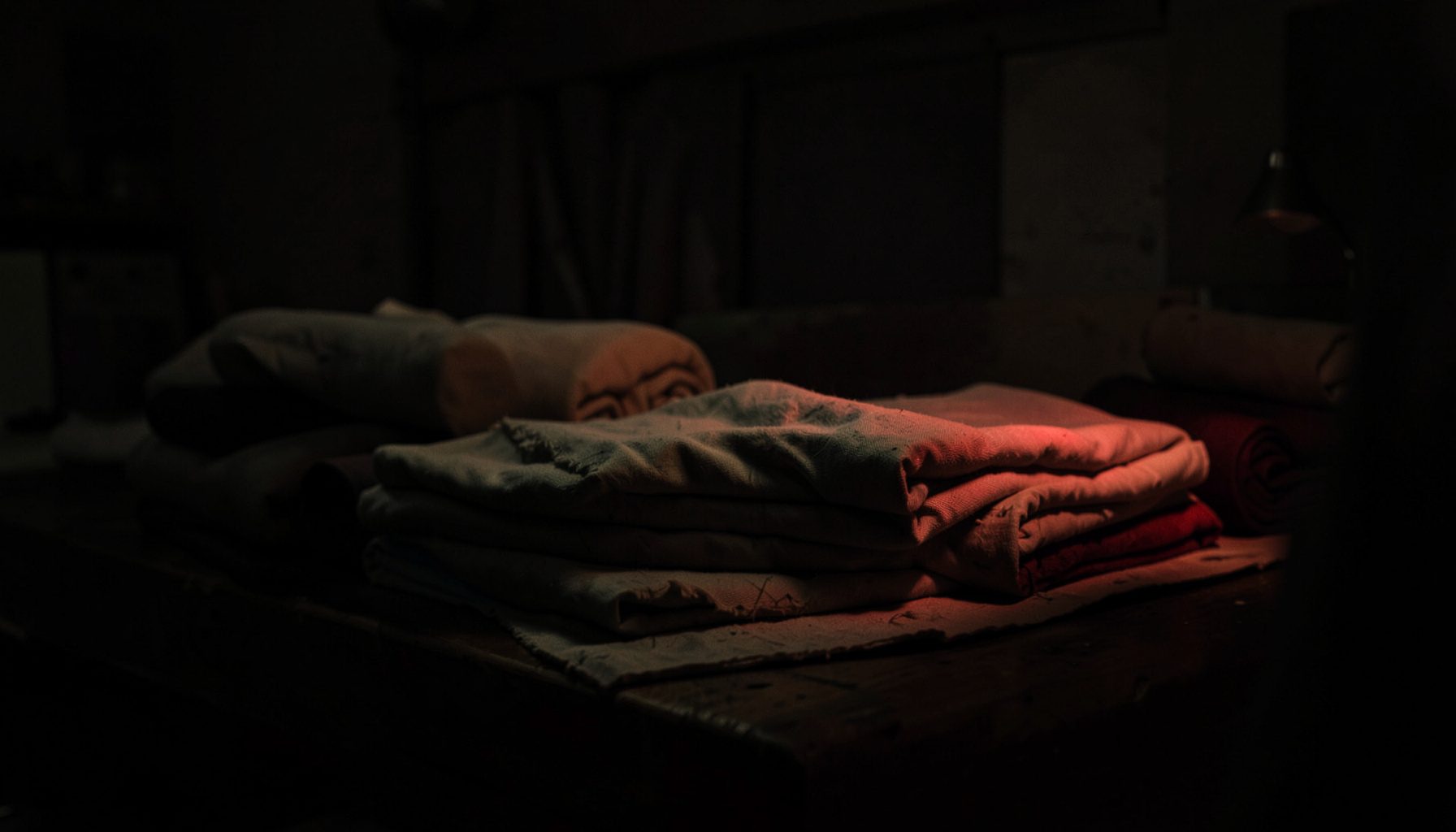 Dark textile workspace with rolls of cotton fabric stacked on a wooden table, deep shadows and warm muted tones